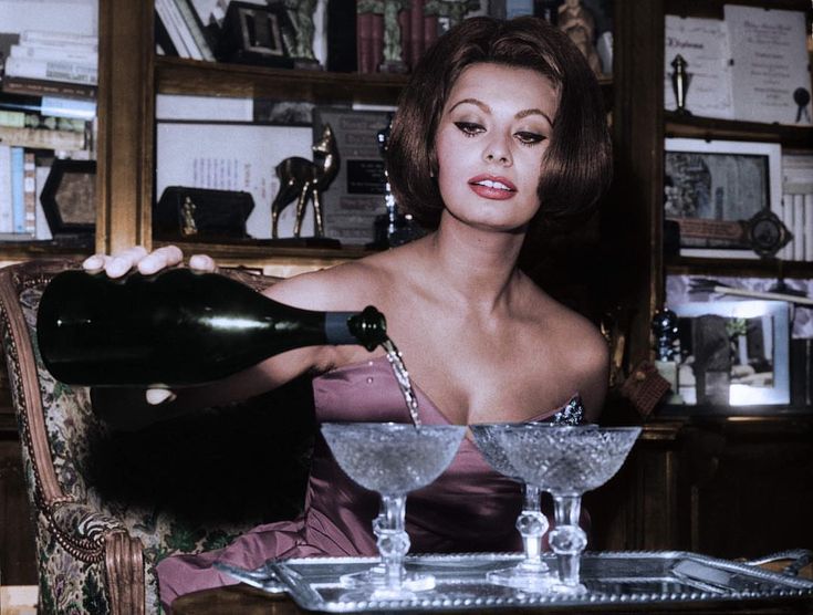 a woman sitting at a table with two martini glasses and a bottle in her hand