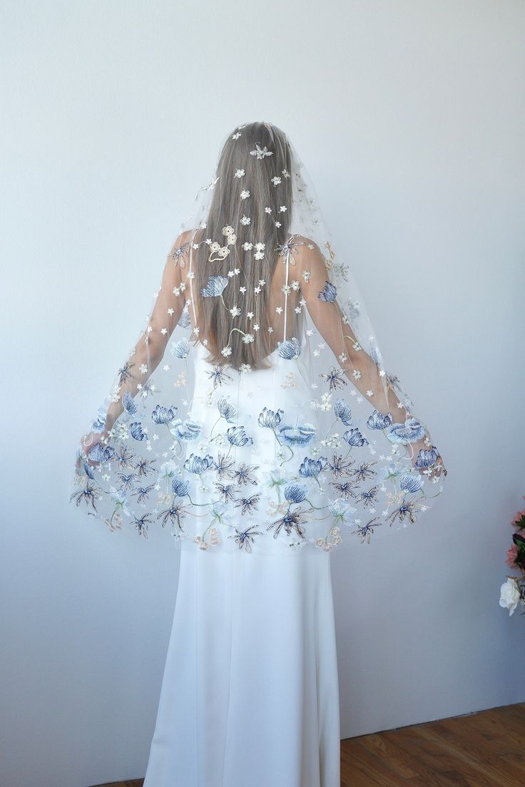 the back of a woman wearing a veil with flowers and butterflies on it, standing in front of a white wall