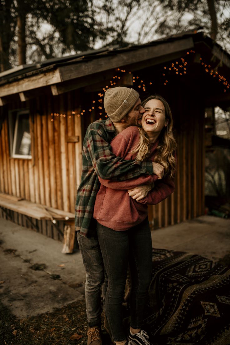 two people hugging each other in front of a wooden cabin with lights on the roof