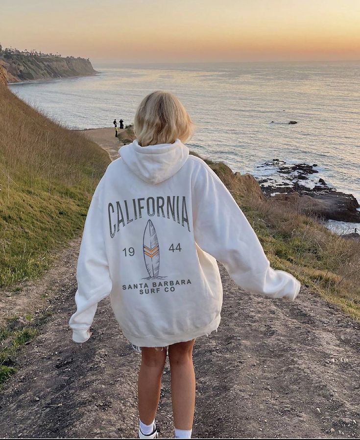 a woman walking down a dirt road towards the ocean with her back to the camera