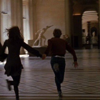 two people are running in the middle of a room with columns and statues behind them