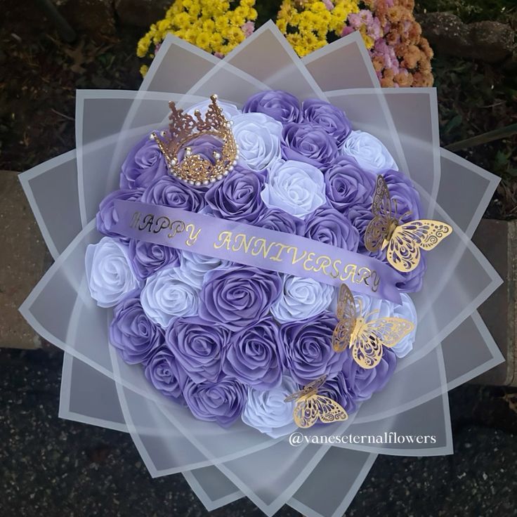 a bouquet of flowers with purple roses and butterflies on it, sitting in front of some flowers