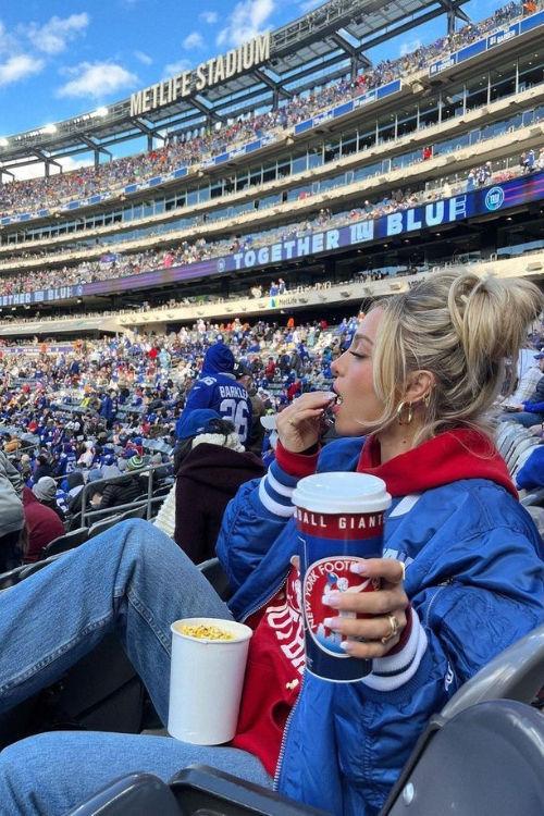 a woman sitting in the stands at a football game eating and drinking from a cup