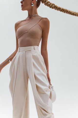 a woman with braids standing in front of a white background wearing beige pants and a one shoulder top