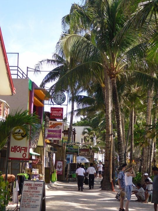 people are walking down the street in front of shops and palm trees on a sunny day