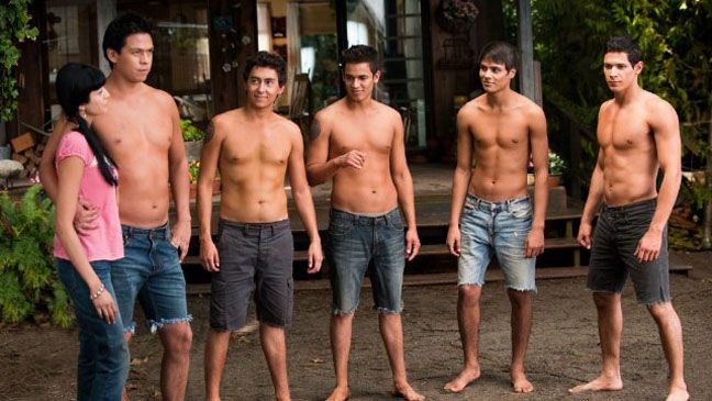 six shirtless men are standing in front of a building and one man is looking at the camera