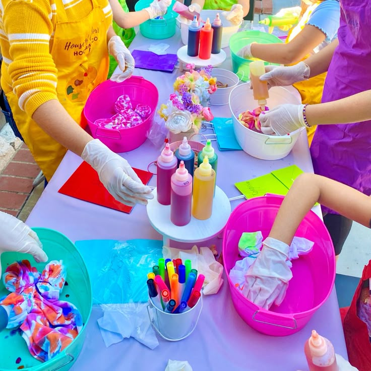 children are making crafts at a table with plastic cups and containers on it, while one child in the background is wearing rubber gloves