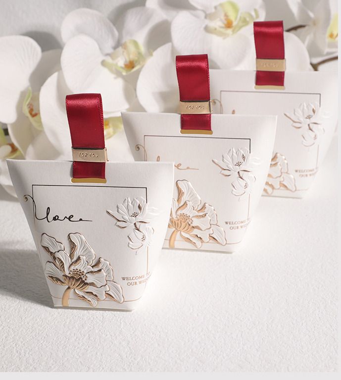 four small white boxes with red ribbons on them and flowers in the background, all lined up