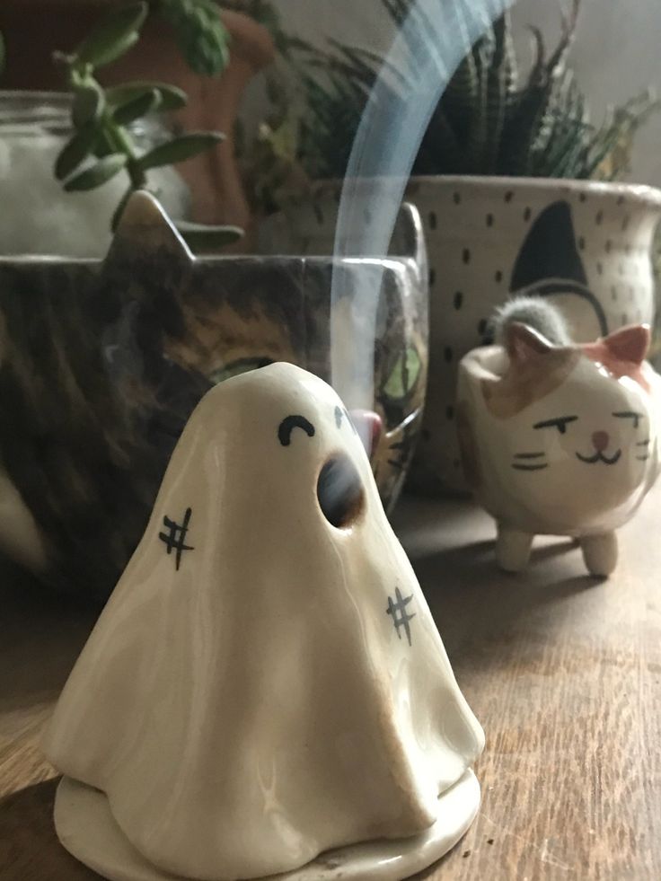 two ceramic ghost cats sitting next to each other on a wooden table with potted plants in the background