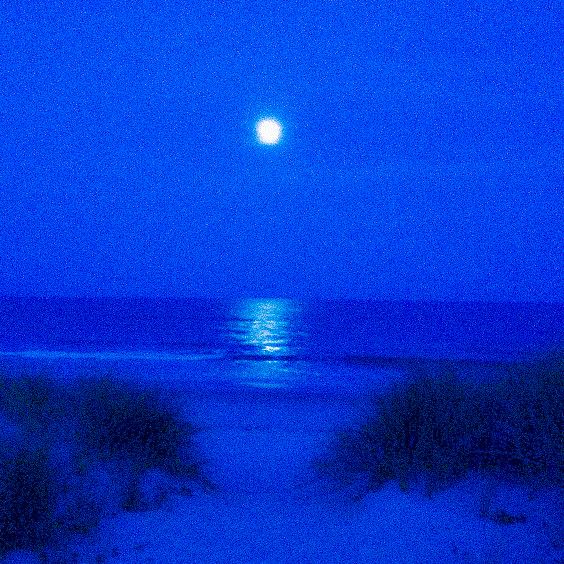 a full moon is seen over the ocean on a cloudy night with blue hues