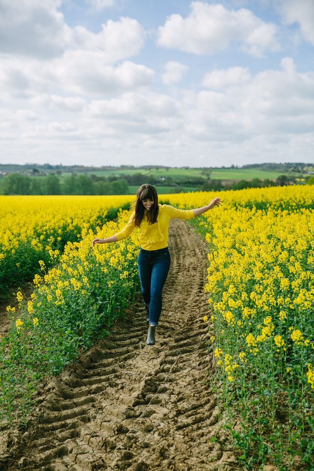 a woman walking down a dirt road in front of a field full of yellow flowers