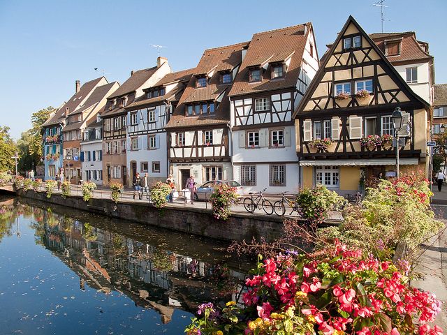 several buildings line the side of a river with flowers in front of them and people walking on the other side