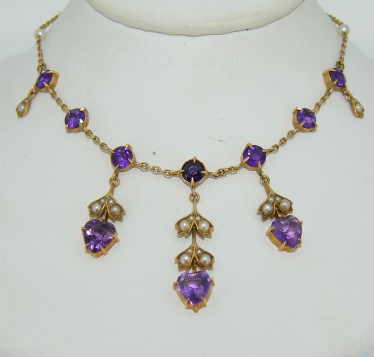 This necklace features fabulous deep purple amethysts. It is crafted from 14K yellow gold, natural amethysts and seed pearls. This necklace dates from the 1900s. It is presented in excellent condition with no repairs or damage. The necklace features three heart drops from the center of the necklace out. The drops encompass a heart cut amethyst as well aa seed pearls adorned on a gold leaf motif. The drops dangle from the body of the necklace. The body features prong set round cut amethysts, two Purple Sapphire Necklace, Victorian Purple Necklace For Anniversary, Antique Purple Necklace For Wedding, Body Features, Crystal Choker Necklace, Leaf Motif, Purple Sapphire, Gold Bead Necklace, Coral And Gold