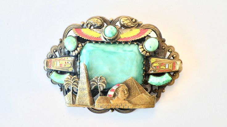 Beautiful and collectible gold filagree Max Neiger Egyptian revival brooch. Large green center glass stone surrounded by Egyptian themed Sphinx, sarcophagi, and pyramids. The paint is in very good condition for age - see photos for close up views. The filagree back shows some slight age related tarnish. The c clasp pin back is functioning and has a slight bend to the pin from use. the brooch measures 2 3/4 by 2 3/4 inches. Max and Norbert Neiger were Czech brothers who produced jewelry in Gablon Collectible Art Deco Carved Jewelry, Art Deco Carved Collectible Jewelry, Ornate Carved Collectible Brooches, Collectible Art Deco Cabochon Brooches, Vintage Carved Collectible Brooches, Art Deco Cabochon Brooches Collectible, Art Deco Antique Gold Collectible Jewelry, Handmade Art Deco Collectible Brooch, Handmade Art Deco Collectible Brooches