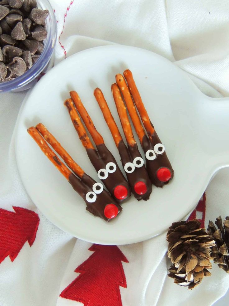 chocolate covered pretzels with googly eyes on a white plate next to pine cones