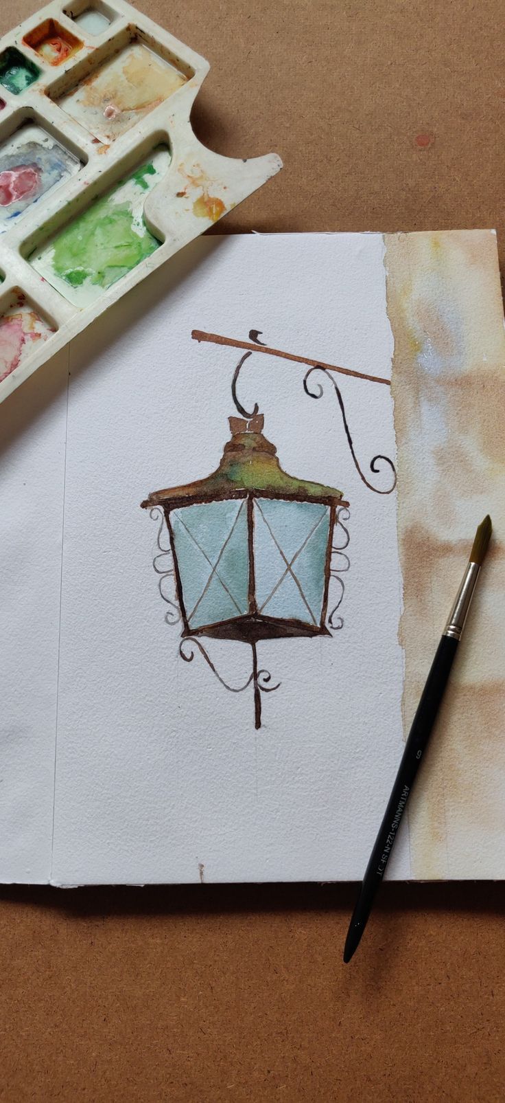 a watercolor sketch of a lamp on paper next to a paintbrush and palette