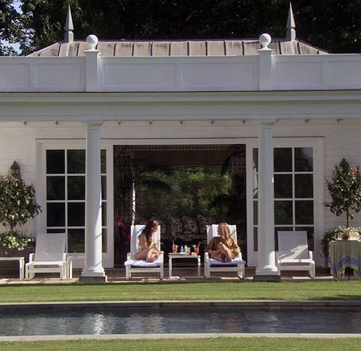 two women lounging on lawn chairs in front of a white house with large windows