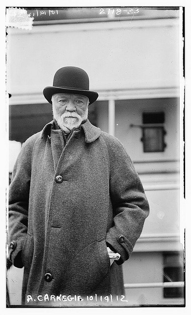 an old man wearing a coat and hat standing in front of a building with his hands in his pockets