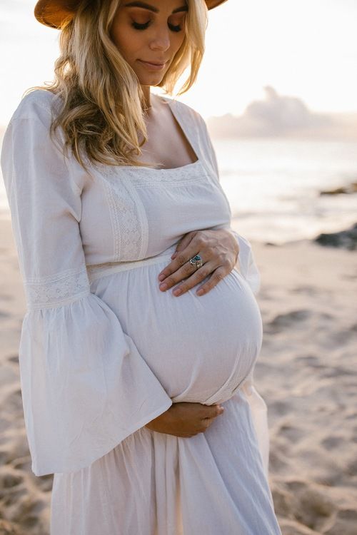 a pregnant woman in a white dress and hat standing on the beach with her belly exposed