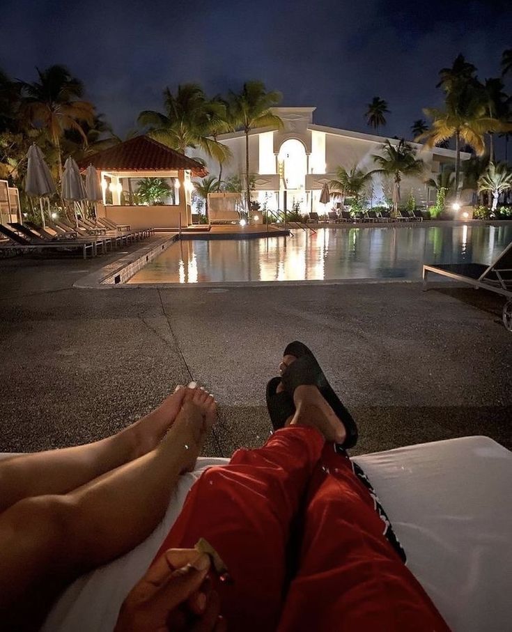 two people laying on the ground in front of a pool at night with their feet up