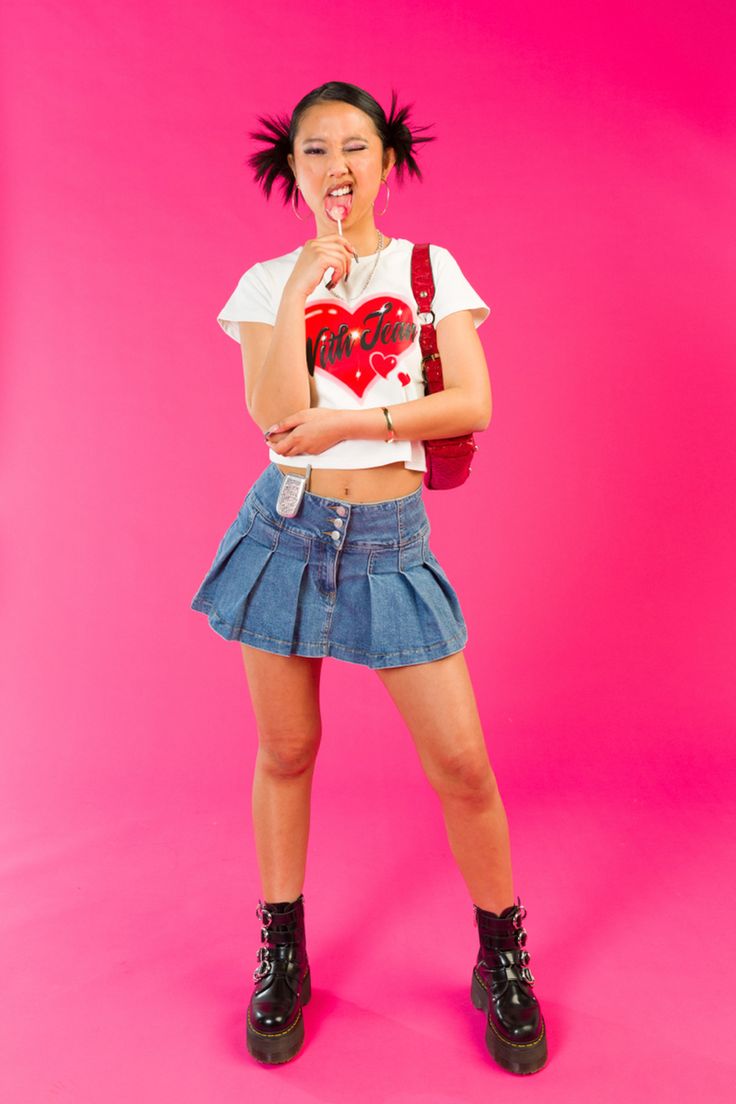 Girl standing in front of a pink backdrop licking a lollipop with her face scrunched up and one eye closed. She is wearing a white with jean top with a red heart on the front, pleated denim skirt, red vintage guess shoulder bag and black platform buckled doc martins. Girly Aesthetic Photoshoot, 2000s Fashion Photography, Y2k Editorial Photoshoot, Y2k Fashion Editorial, Y2k Inspired Photoshoot, Y2k Fashion Photoshoot, 2000s Fashion Photoshoot, Early 2000 Photoshoot, 2000 Fashion Women