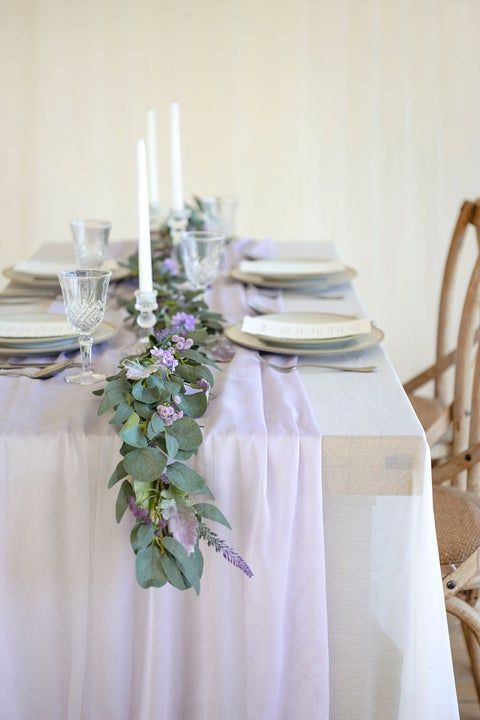 the table is set with white plates and silverware, purple flowers and greenery