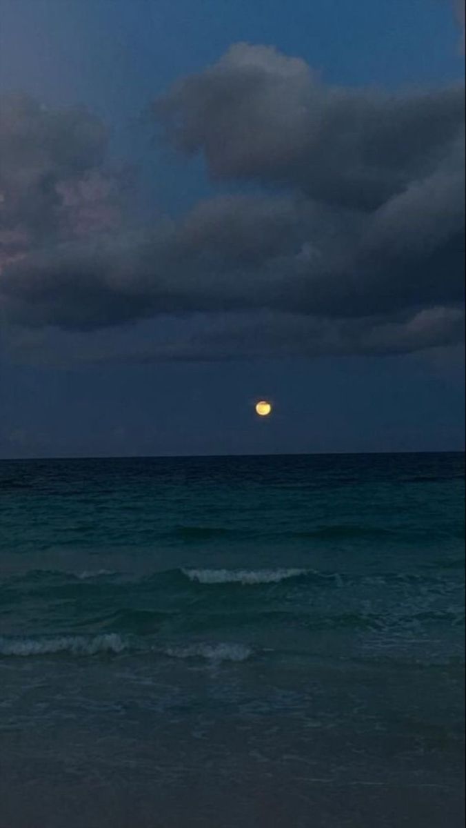 the moon is setting over the ocean with dark clouds