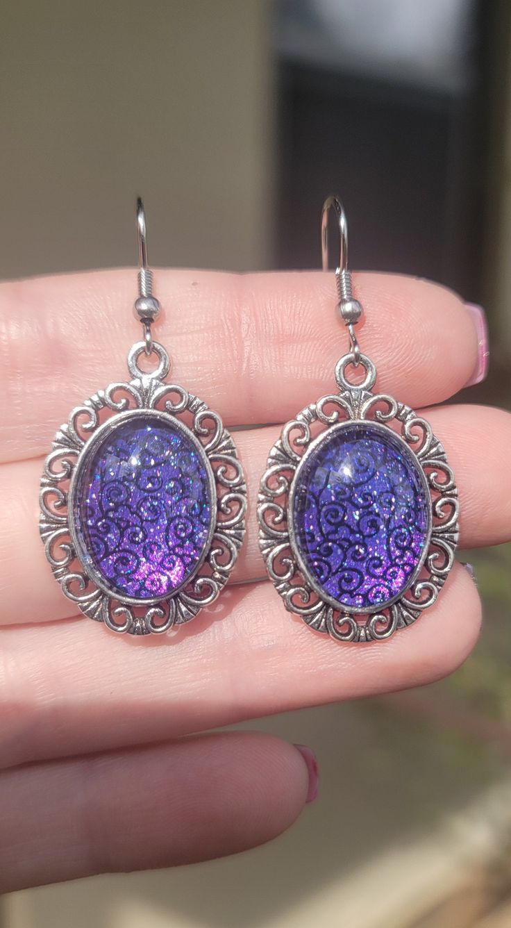 The design in these earrings was hand created with nail polish!  This pair features a glittery purple base with a black design.  The design is set with glass in a silver metal alloy base, and features hypoallergenic 316 Surgical Steel ear wires.  Inner Diameter/Design Size: 13mmx18mm Outer Diameter: 22mmx30mm Total Length with Ear Wire: 50mm Silver Glitter Earrings For Party, Silver Resin Earrings For Party, Purple Crystal Jeweled Earrings, Purple Resin Jewelry For Party, Purple Drop Crystal Earrings, Purple Dangle Crystal Earrings, Silver Glitter Round Earrings, Purple Glitter Party Jewelry, Purple Resin Party Jewelry