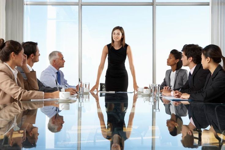 a woman standing in front of a group of people sitting at a table with their hands together