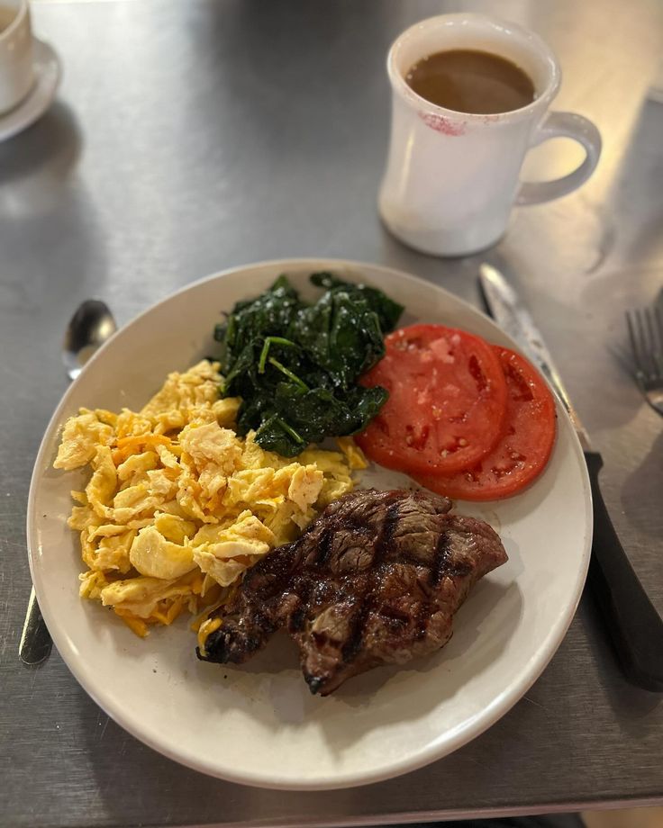 a white plate topped with meat, eggs and vegetables next to a cup of coffee