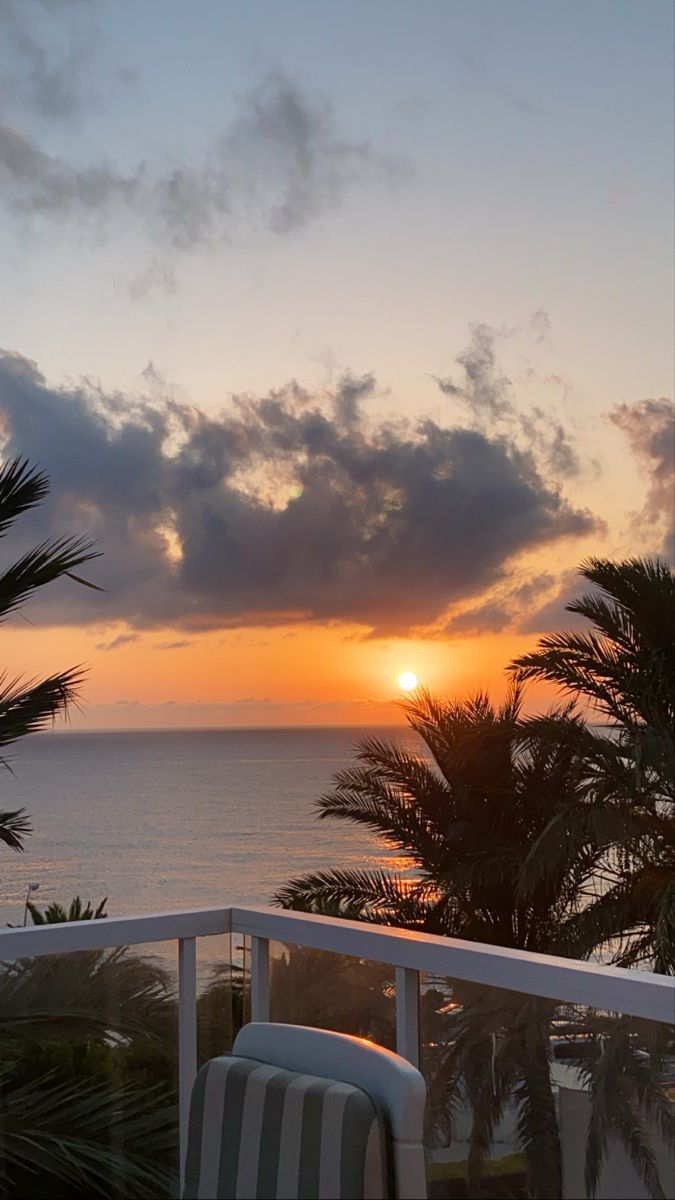 the sun is setting over the ocean with palm trees in front of it and a chair on the balcony