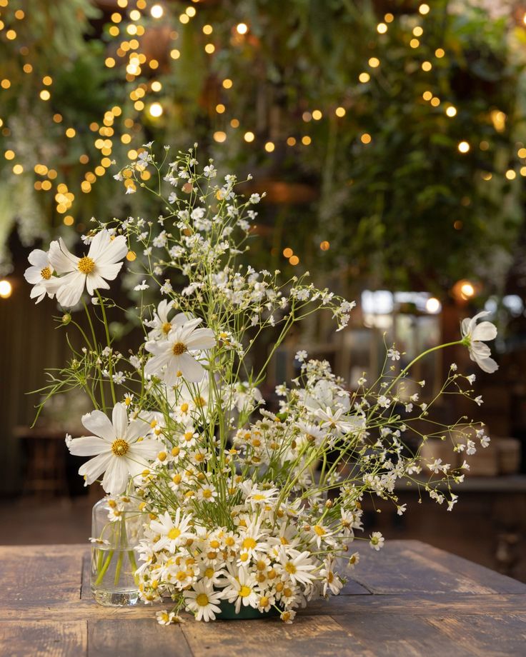 a vase filled with white flowers sitting on top of a wooden table covered in lights