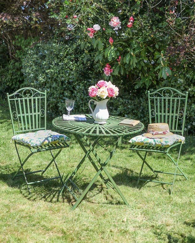 a table and chairs in the grass with flowers on it, near a bush full of pink roses