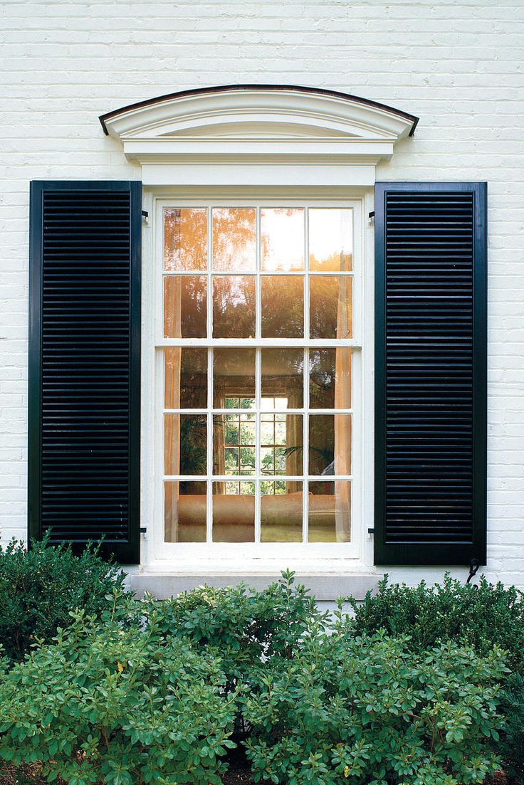 an open window with black shutters on a white brick wall and shrubbery in the foreground