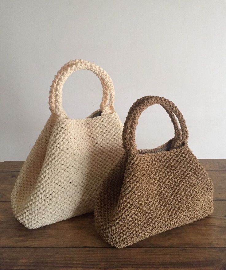 two straw bags sitting on top of a wooden table next to each other, one is white and the other has brown handles