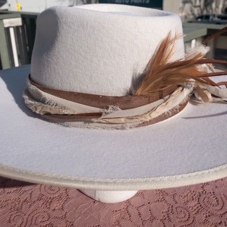 You will always be ready to go and styled with this gorgeous hatband on your hat. The band is 22.5" x 1.5" and has adjustable ties in the back so it can fit hats from 22.5-34". The bottom layer is one of our favorite leathers, a metallic toasted bronze pink. Very subtle but gorgeous! The designer has then used two types of silk ribbons that complement the leather and really bring the color to life. The brown suede lace adds a nice complete to complete the top layer. Beautiful accent feather comp Ribbon Hat Bands, Luxury Brown Hat Bands For Country Events, Luxury Adjustable Cream Hat Band, Luxury White Country Hat Bands, Flea Style Hat Bar, How To Make A Hat Band, Custom Hat Bar Ideas, Hat Band Ideas, Diy Hatbands