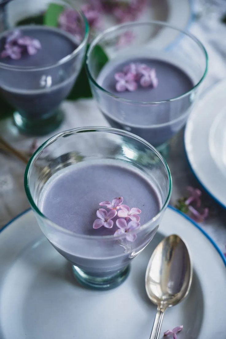 two cups filled with liquid and flowers on top of white plates next to silver spoons