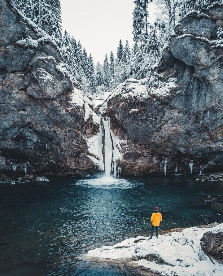 a man standing on top of a snow covered rock next to a waterfall in the middle of a forest