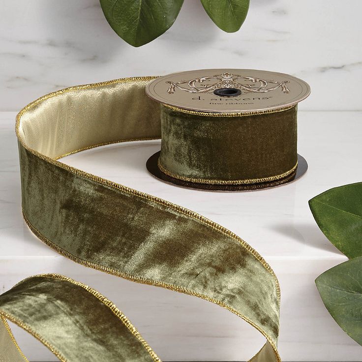 a roll of green velvet ribbon with gold trim on a white table next to leaves