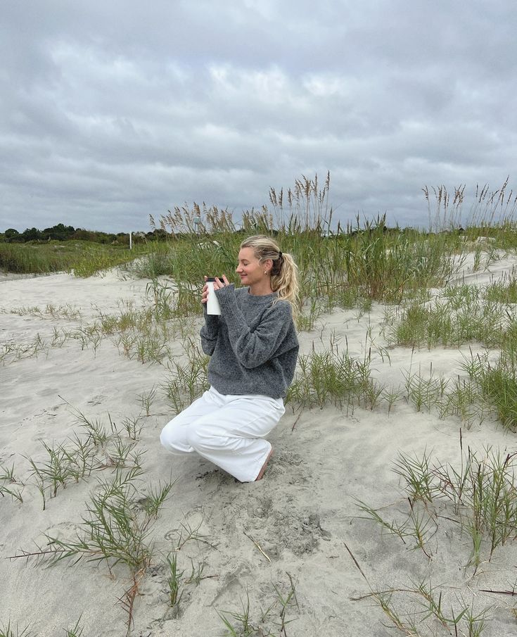 a woman kneeling in the sand drinking from a water bottle while wearing white pants and a gray sweater