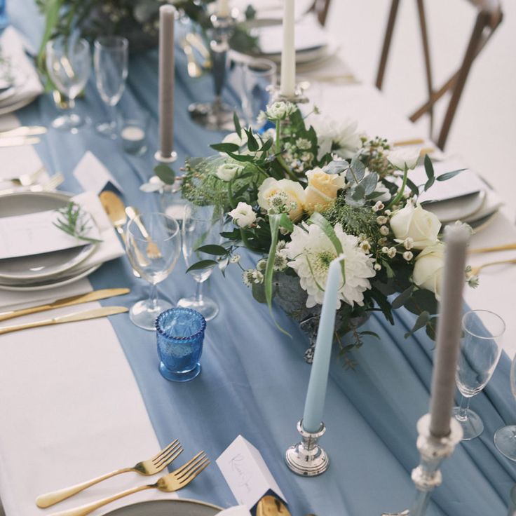 the table is set with white and blue flowers, silverware, and gold place settings