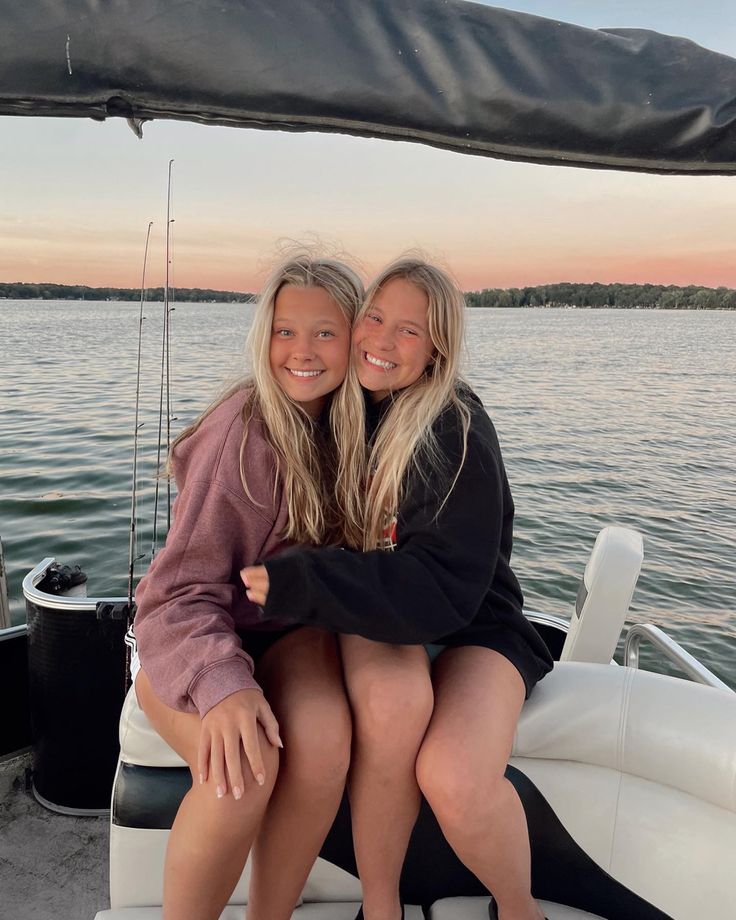 two women sitting on the back of a boat in the water at sunset, hugging each other