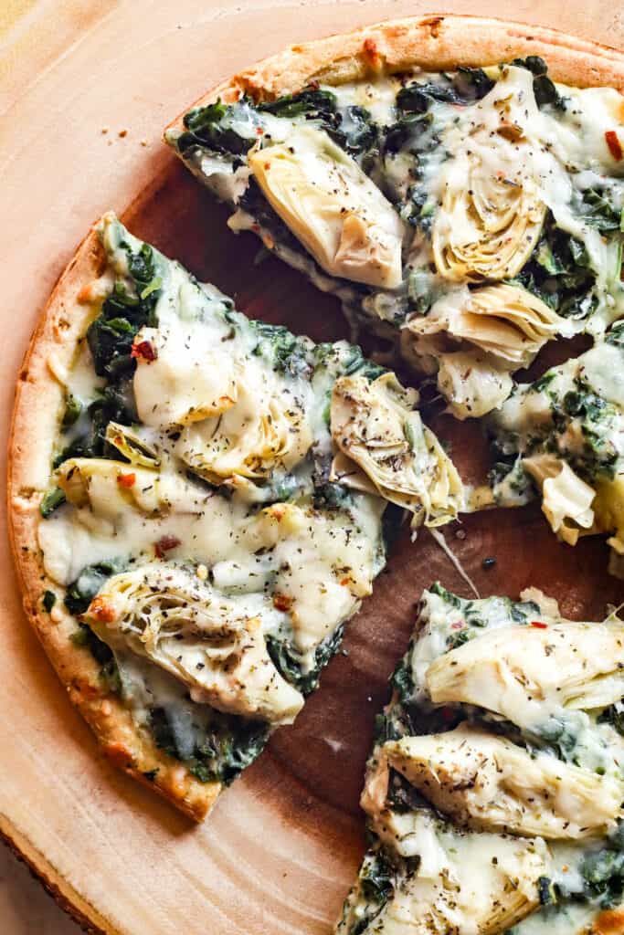 a pizza with spinach, artichokes and cheese is on a wooden platter