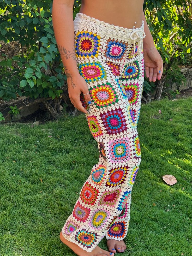 a woman standing in the grass wearing colorful crochet pants and sandals with her hand on her hip