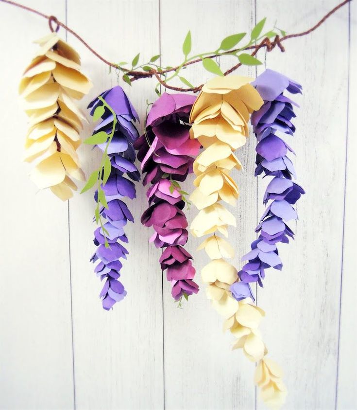 paper flowers are hanging from a branch on a white wooden wall, with green leaves in the foreground