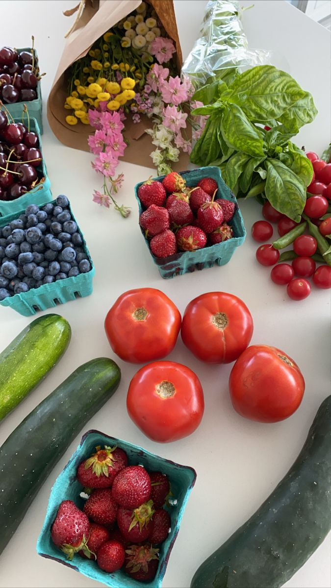 various fruits and vegetables are arranged on a white counter top, including cucumbers, strawberries, tomatoes, blueberries, green beans, and more