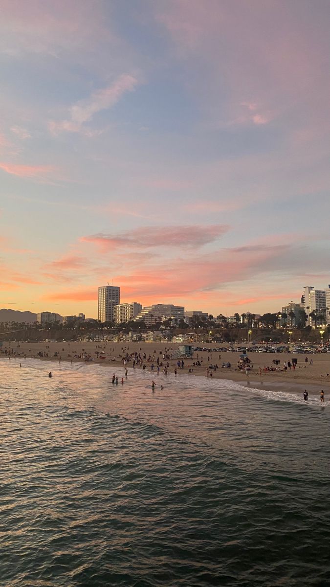 people are on the beach at sunset with buildings in the backgroung and pink clouds