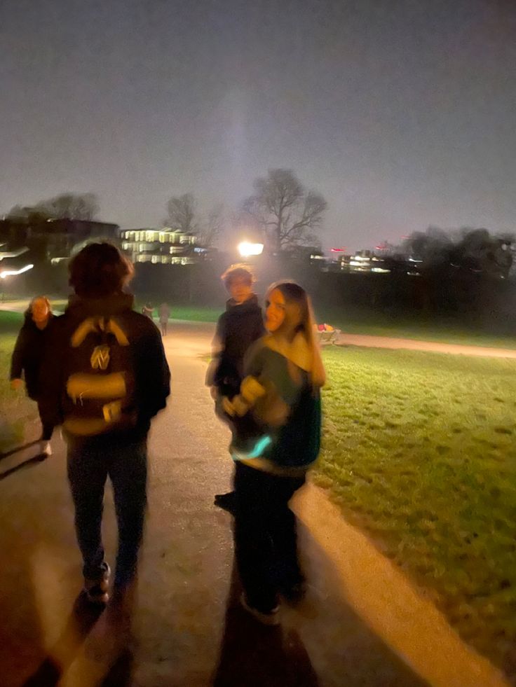 three people walking down a path at night with lights on in the back ground and grass behind them