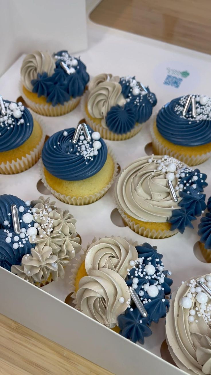 twelve cupcakes with blue and white frosting are in a box on the table