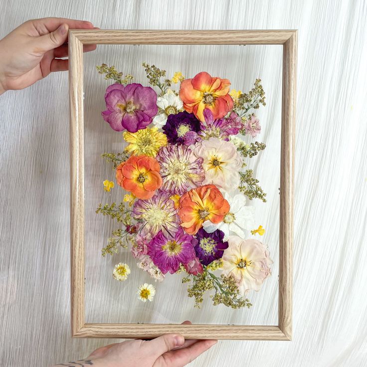 a person holding a frame with flowers in it on a white tableclothed surface